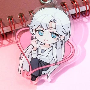 以下が含まれることがあります： 白い髪と青い目のアニメ風のキャラクターが、ピンクのハートの輪郭の中に座っている透明なアクリルキーホルダー。キーホルダーには、銀色のハート型の留め金が付いています。キャラクターは白いトップスと濃い色のパンツを着用しています。