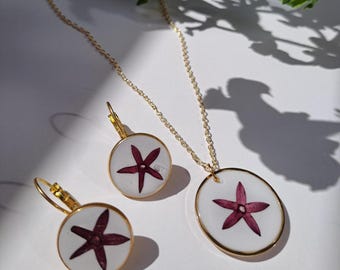 Puede incluir: Un conjunto de joyas doradas que incluye un collar y pendientes. Cada pieza tiene un fondo blanco con un diseño floral en forma de estrella de color granate. Los pendientes son redondos y el colgante del collar es ovalado.