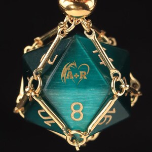 May include: A gold-toned necklace featuring a teal, twenty-sided die pendant encased in a delicate gold metal cage. The die displays the number 8 and a custom engraving of the letters A+R inside a heart shape, suspended from a matching chain.