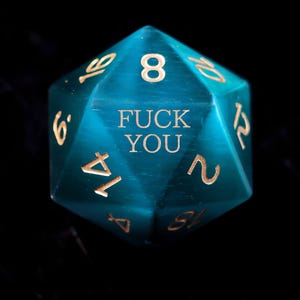 Könnte beinhalten: Ein türkisblauer, vielseitiger Würfel mit goldfarbenen Zahlen und den Worten "FUCK YOU" auf einer Seite. Der Würfel befindet sich vor einem dunklen, reflektierenden Hintergrund.