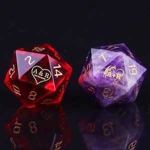May include: Two twenty-sided dice. One is transparent red with gold numbers and the text "A & R 03/14/2030" inside a heart. The other is purple with gold numbers and a similar heart design. Both dice are on a reflective black surface.