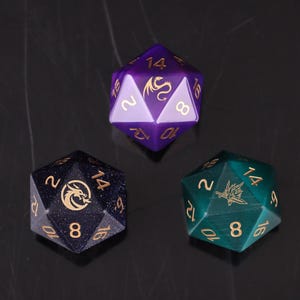 May include: Three twenty-sided dice featuring gold-colored numbers and dragon icons. One die is purple with lighter purple accents, one is dark blue with glittery flecks, and one is a deep teal with a cat-eye effect finish on the surface.