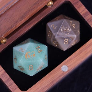 May include: Two twenty-sided dice resting in a wooden box. One die is light green with gold numbers and the engraving A+R inside a heart. The other die is a muted grey-brown with gold numbers and the engraving S&M 01/31/26 inside a floral heart.