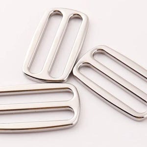 May include: Three shiny, silver-colored metal slider buckles. Each buckle has a rectangular shape with two parallel bars running across the center, creating three openings. The buckles are likely used for straps or belts.