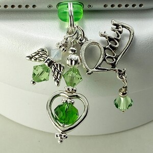 Peut inclure: Un porte-téléphone argenté avec des détails verts. Le charme comprend un pendentif en forme de cœur avec un cristal vert, des ailes d'ange et le mot "love" dans une police décorative. D'autres cristaux verts et perles argentées complètent le design.