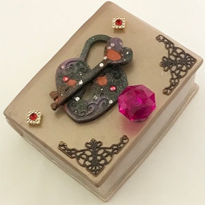 May include: A decorative book-shaped box with a heart-shaped lock and key. The box is beige with ornate metal corner accents and red jewel embellishments. A large, faceted pink gem sits on the side.