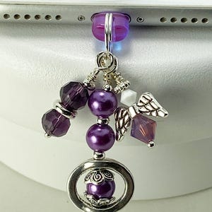 May include: A purple phone charm with a silver ring and angel wing charm. The charm features various purple beads, including faceted and round beads, and a small white cube. The charm is attached to a purple plug.