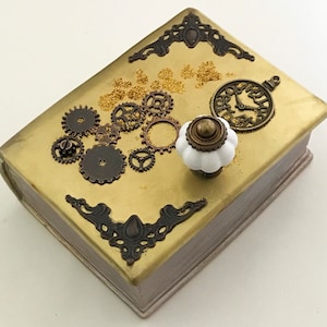 May include: A decorative box with a gold-painted surface, adorned with bronze-colored gears, corner accents, and a clock-shaped charm. It features a white and bronze knob. The box has a steampunk theme.