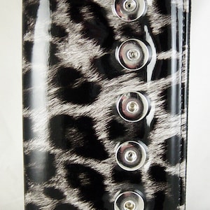 May include: A black and white leopard print wallet with a glossy finish. The wallet has a silver zipper and five silver snap closures down the front. The design resembles animal fur.
