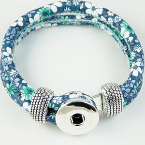 May include: A fabric bracelet with a floral pattern in shades of blue, green, and white. The bracelet has a silver snap closure and decorative silver beads on each side of the clasp. The bracelet is a wrap style.