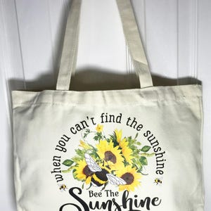 May include: Cream-colored tote bag with a floral and bee design. The bag features a bouquet of yellow sunflowers and a bumblebee, with the text "When you can't find the sunshine, Bee The Sunshine."
