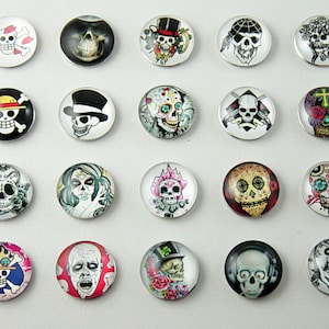 May include: A collection of twenty-one round glass cabochons featuring various skull designs. The skulls are decorated with hats, flowers, and other embellishments. Some designs include crossbones and pirate-themed imagery. The cabochons are set against a white background.