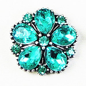 May include: A flower-shaped brooch with a silver-tone metal base. The brooch features five large, teardrop-shaped, teal-colored crystals surrounded by smaller, round teal crystals. The crystals are set in a detailed, ornate design.