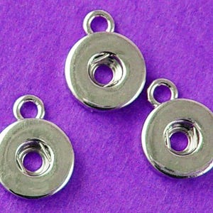 May include: Three silver-colored snap fasteners with a loop at the top for attaching to fabric. The fasteners are round with a central hole and are displayed against a purple background.