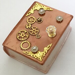 May include: A steampunk-themed box resembling a book. The copper-colored top is adorned with gold-colored gears, floral corner accents, and pearl-centered embellishments. A clear, faceted knob with a gold base serves as a handle.