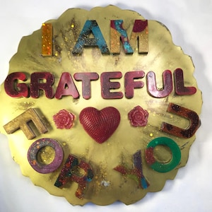 May include: A gold-colored resin art piece with the words "I AM GRATEFUL FOR YOU" in colorful, textured letters. The piece includes a red heart and two rose shapes. Glitter accents the surface.
