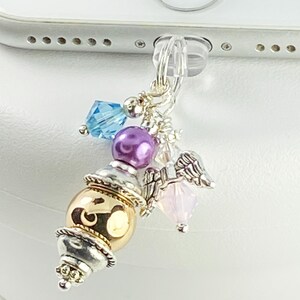 May include: A silver phone charm with a decorative design. The charm features a gold and silver bead, a purple bead, a blue crystal, a pink crystal, and small silver angel wings. The charm is attached to a clear plastic plug.