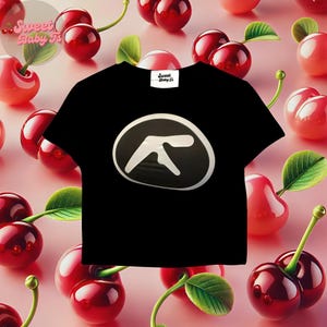 May include: Black t-shirt with a white logo of a stylized letter A on a black circle. The shirt is surrounded by red cherries on a pink background. The text "Sweet Baby T's" is on a white tag attached to the shirt.