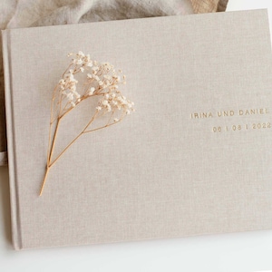 May include: A beige linen-covered photo album with gold lettering that reads "Irina und Daniel 06.08.2022". A sprig of white baby's breath is placed on the front cover.