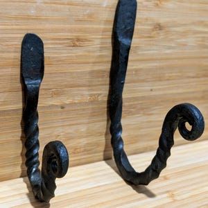 May include: Two black, hand-forged metal hooks. The hooks have a twisted design on the lower portion and a curled end. The hooks are mounted on a light brown wooden surface.