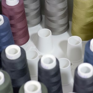 May include: A selection of thread spools in various colours, including green, blue, black, brown and grey, are displayed in a white plastic holder. The spools are all of the same size and shape, and each is wound with thread.