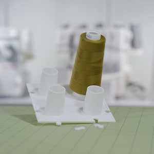 May include: A white plastic spool holder with three spools, one of which is filled with olive green thread.