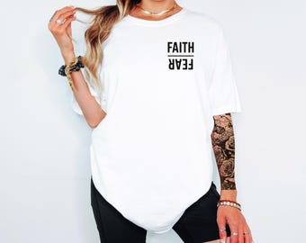 Puede incluir: Una camiseta blanca de manga corta con la palabra FAITH impresa en negro sobre una línea horizontal, y la palabra FEAR impresa en negro al revés debajo de la línea en la parte superior izquierda del pecho de la prenda.