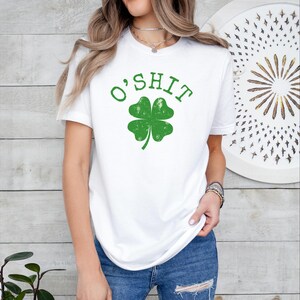Puede incluir: Camiseta blanca con el texto "O'SHIT" sobre un trébol verde. El gráfico tiene un aspecto desgastado. La camiseta se combina con unos vaqueros azules.