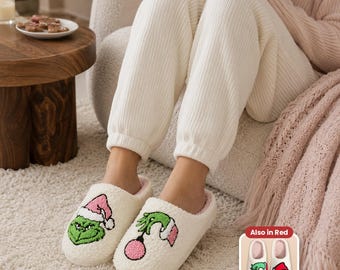 Può includere: Pantofole in peluche bianco sporco con un personaggio verde con un cappello da Babbo Natale rosa a sinistra e una mano verde che tiene un ornamento rosa a destra. Un riquadro mostra le stesse pantofole in rosso e bianco con il testo Also in Red.