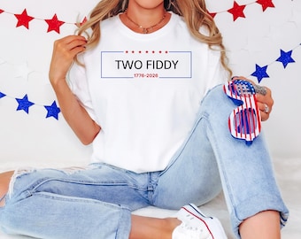May include: A white short-sleeve t-shirt featuring a blue rectangular border with six red stars above the text TWO FIDDY and the years 1776-2026 below. The shirt is paired with light blue distressed denim jeans and red, white, and blue shutter glasses.