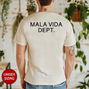May include: Beige unisex t-shirt with the text "MALA VIDA DEPT." printed in black. The shirt has short sleeves and a crew neck. A red circle with the words "UNISEX SIZING" is visible in the lower left corner.