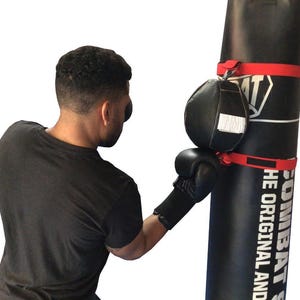 May include: A person wearing black boxing gloves punches a black freestanding punching bag with red straps. The bag has the text "COMBAT SPORTS THE ORIGINAL AND" printed on it.