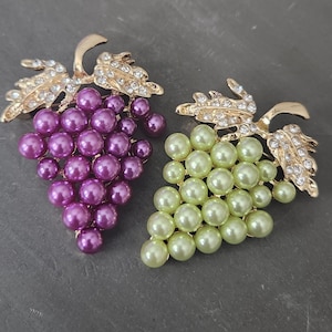 May include: Two grape-shaped brooches. One brooch features purple pearl-like beads, while the other has green pearl-like beads. Both have gold-colored leaves with clear crystal accents. These brooches are perfect for adding a touch of elegance.