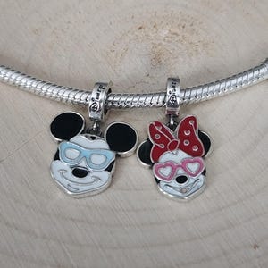 May include: Two silver-toned Mickey Mouse and Minnie Mouse charm pendants. Mickey wears light blue sunglasses, and Minnie has a red bow and pink heart-shaped glasses. Both charms have black ears and are attached to a silver chain.