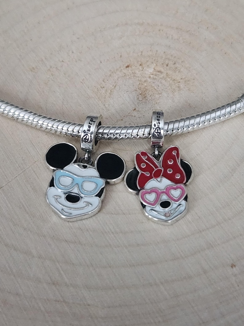 May include: Two silver-toned Mickey Mouse and Minnie Mouse charm pendants. Mickey wears light blue sunglasses, and Minnie has a red bow and pink heart-shaped glasses. Both charms have black ears and are attached to a silver chain.