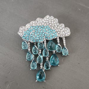 May include: A silver-toned brooch in the shape of a cloud with sparkling clear and turquoise blue crystals. Teardrop-shaped blue crystals hang from the cloud, creating a rain effect. The brooch is set against a gray background.