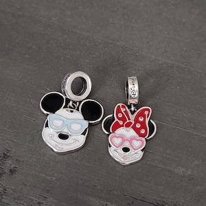 May include: Two silver charms featuring cartoon mouse faces. One has blue glasses, the other has a red bow with white polka dots and pink heart-shaped glasses. Both have black ears and white faces.