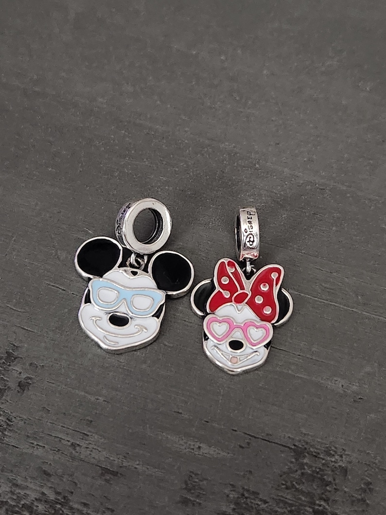 May include: Two silver charms featuring cartoon mouse faces. One has blue glasses, the other has a red bow with white polka dots and pink heart-shaped glasses. Both have black ears and white faces.
