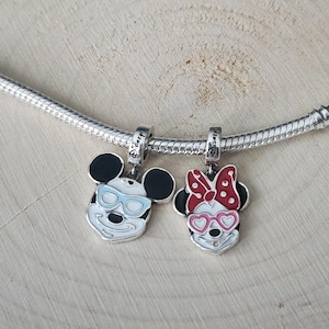 May include: Two silver charms featuring Mickey and Minnie Mouse. Mickey has black ears, a white face, and blue glasses. Minnie has black ears, a white face, and pink heart-shaped glasses and a red bow. Both charms are on a silver chain.