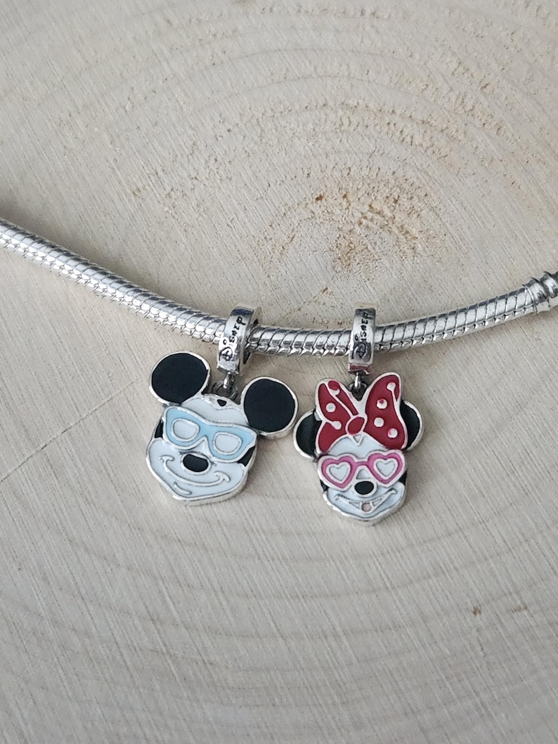 May include: Two silver charms featuring Mickey and Minnie Mouse. Mickey has black ears, a white face, and blue glasses. Minnie has black ears, a white face, and pink heart-shaped glasses and a red bow. Both charms are on a silver chain.