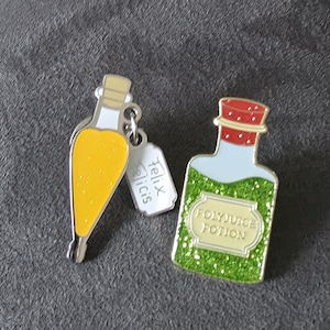 May include: Two metal enamel pins. One is a yellow teardrop-shaped bottle with a small white tag labeled Felix Felicis. The other is a rectangular bottle with a red cap and green glittery liquid, featuring a label that reads POLYJUICE POTION.