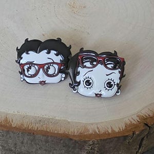 May include: Two enamel pins featuring the head of a cartoon character with black hair and white skin. One pin shows the character wearing red glasses, while the other pin shows the character with red glasses resting on top of their black hair.