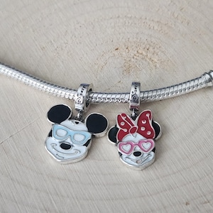 May include: Two silver charms featuring Mickey and Minnie Mouse. Mickey has blue sunglasses, and Minnie has a red bow and pink heart-shaped glasses. Both charms have black ears and are on a silver chain.