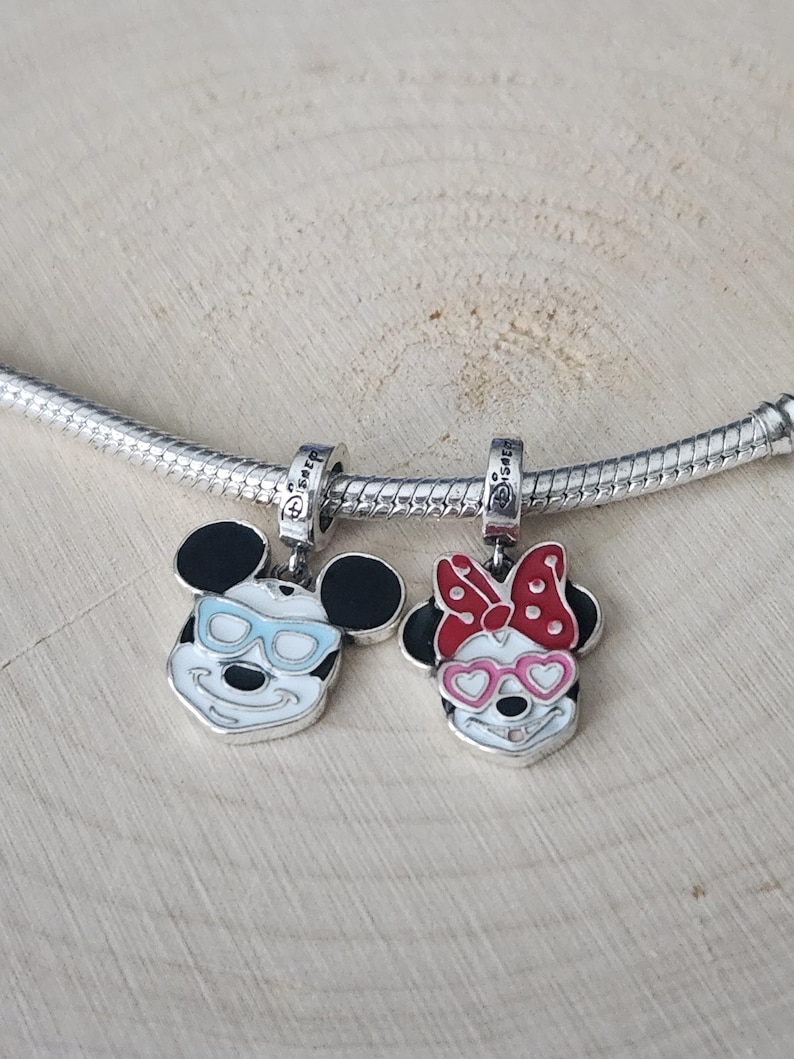 May include: Two silver charms featuring Mickey and Minnie Mouse. Mickey has blue sunglasses, and Minnie has a red bow and pink heart-shaped glasses. Both charms have black ears and are on a silver chain.