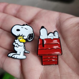 May include: Two enamel pins featuring Snoopy from the Peanuts comic strip. One pin shows Snoopy with Woodstock, the other shows Snoopy on top of his red doghouse. Both pins have black outlines and bright colors.