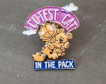 May include: An enamel pin featuring an orange cartoon cat holding a small tan teddy bear. Above the cat is a purple arch with white clouds and the text CUTEST CAT. Below the cat is a blue rectangular banner with the text IN THE PACK.