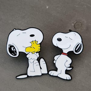 May include: Two Snoopy character pins. One pin shows Snoopy sitting and holding Woodstock, the yellow bird. The other pin shows Snoopy standing with his hands behind his back. Both pins have a white body, black accents, and a black outline.