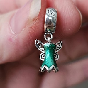 May include: A silver charm with a green and white fairy design. The fairy has detailed wings and a dress with a textured, glittery finish. The charm has a loop at the top with an engraved design.