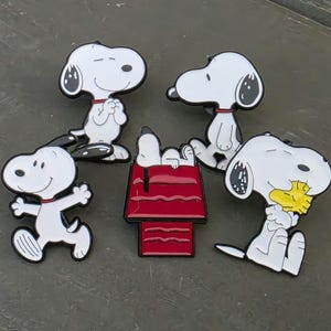 May include: A collection of five enamel pins featuring the white cartoon dog Snoopy in various poses, including sitting, running, and hugging a yellow bird, alongside a red doghouse with a black outline and white Snoopy figure on top.