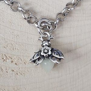 May include: A silver-toned necklace featuring a detailed bee pendant. The bee pendant has openwork wings and a small, light green teardrop-shaped stone. The necklace has a chunky chain and a toggle clasp.